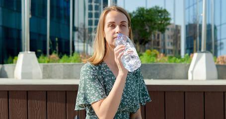 Young student woman sitting near modern buildings and drinking water