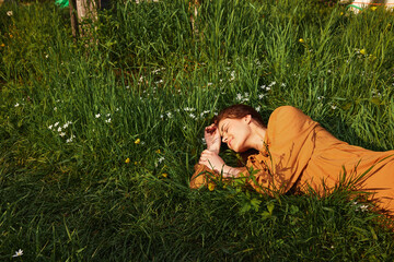 horizontal photo of a happy woman lying in the grass in a long orange dress, with her eyes closed and a pleasant smile on her face, with her hand behind her head.