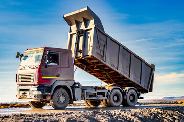 Dump truck with a raised body at a construction site. The process of transportation and unloading of soil. Technique for transportation of bulky materials. Rental of construction equipment. - obrazy, fototapety, plakaty