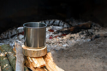 A metal mug for camping stands on firewood by the fire. Camping dishes, fire, evening. Copyspace