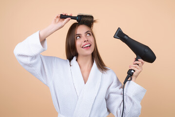 Woman in bathrobe combing hair, drying hairs with hairdryer in studio. Portrait of female model...