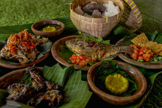 Traditional sasak dish consisting of ayam merangkat, fried tilapia, fried chicken, fried tempe with sambal, clear vegetable soup, and rice served on a clay plate and banana leaves.