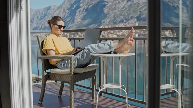 A young Caucasian woman working on a laptop while sitting on the terrace with a beautiful sea view on a summer sunny day.