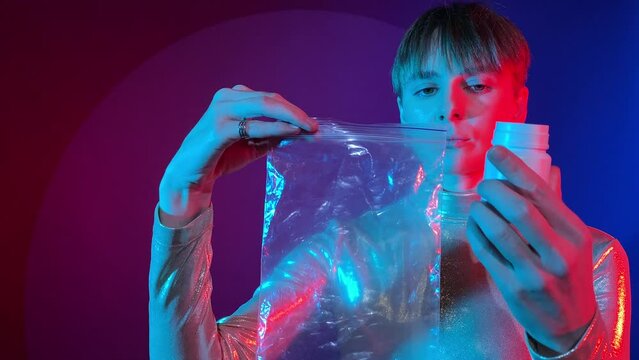 Serious young woman blank expression pouring pills out packer bottle into plastic bag neon red blue lighting on colorful background. Female detective criminalist taking drugs as evidence. Social issue
