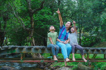 Happy young mother with little girls sitting on wooden bridge near river in summer. Mother and two daughters having fun while spending time together outdoors. Happy loving family and Mother's Day.