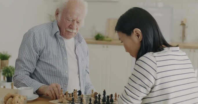 Retired man playing chess with young woman volunteer enjoying game and talking in kitchen at home. Entertainment and retirement concept.