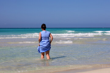 Fototapeta na wymiar Woman in blue dress going to azure sea water. Travel and beach vacation on sandy coast