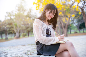 Portrait cute smiles Asian of attractive young teenage girl a holding diary book writing note at Yellow Tabebuia Chrysotricha flowers with the park in spring day at Evening background in Thailand.