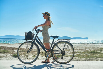 Carefree woman with bike riding on sand beach having fun, on the seaside promenade on a summer day.
