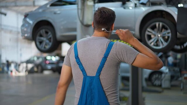 Back view thoughtful unsure Caucasian man in uniform scratching head looking at broken vehicle in car service station. Hesitating expert repair man thinking planning car maintenance. Slow motion