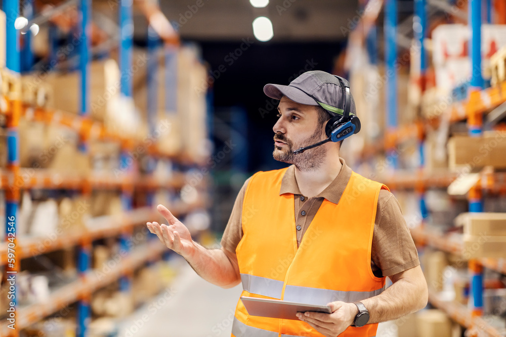 Wall mural A delivery center manager is talking on headset with workers while standing in storage with tablet.