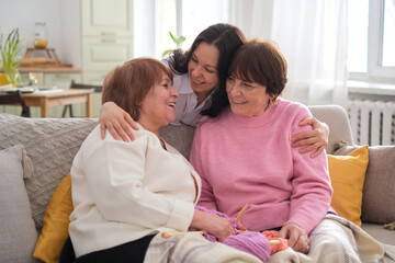 Three joyful women from distinct generations engage in heartfelt conversations, hugs, and laughter in a comfortable living space.role of strong family connections in mental health and well-being.