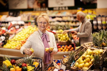 A happy senior woman is purchasing fresh fruits at the supermarket while smiling at the camera.