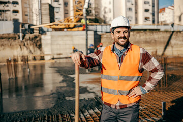 A happy site worker is leaning on shovel and smiling at the camera on site during concreting...
