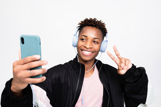 Afro american young hipster wearing new modern wireless headphones holding cell phone smartphone in hands listening to music enjoying songs taking selfie picture shwoing two fingers at camera.