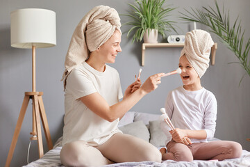 Indoor shot of smiling joyful cheerful mother and daughter in towels on bed at home having spa beauty day mommy applying powder kid;s nose smiling happily.