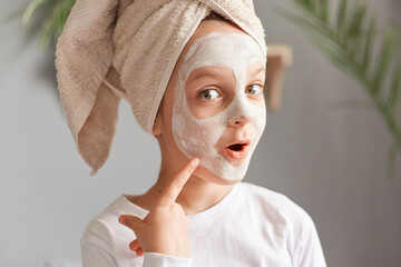 Astonished little girl with towel on head with cosmetic mask pointing at her face with excited look doing skin care procedures wearing white shirt.