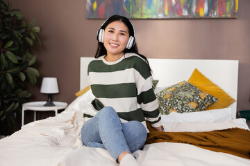 Kazachstan delighted pretty beautiful woman in modern new laste series headphones girl wearing casual outfit clothes looking at camera smiling sitting laying on a bed in bedroom.