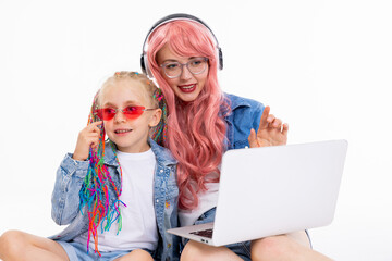 Young mom with pink wig and new headphones and glasses pointing at laptop watching cartoons movie with curious daughter with colorful braids and pink sunglasses. Girls wearing denim clothes.