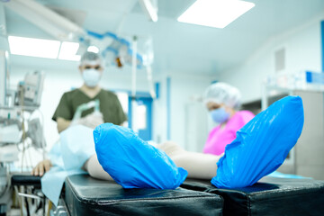 Selective focus on the foot of a person lying on the operating table. Blue medical cellophane shoe covers on the patient's feet against the background of blurred silhouettes of doctors.