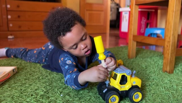 Cute and expresive 3-year-old black child playing at home to fix his yellow toy bulldozer laying on a green rug.