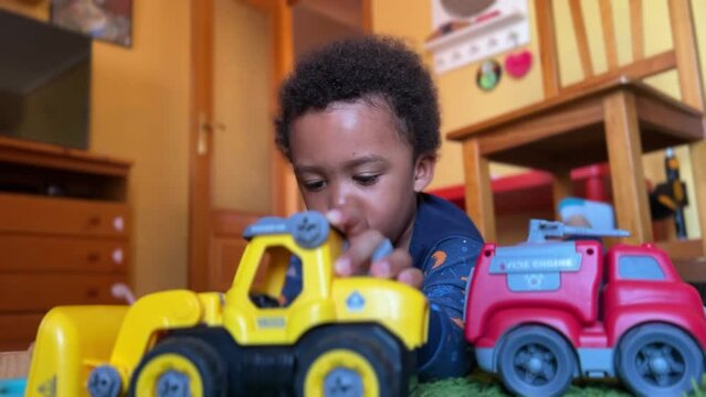 Scene of a cute and expressive  3-year-old black child playing at home to fix his toys laying on the a red rug and wearing a blue pyijamas.