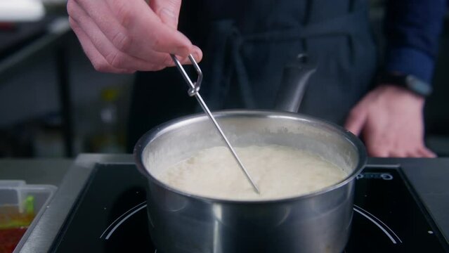 Close up shot of chef cook mixing cream sauce by spiral whisk in stewpan on electric stove. Cook prepares food for guests. Process of making delicious dishes. Work on restaurant kitchen. Slow motion.