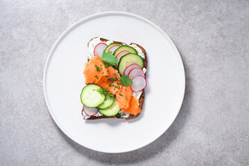 Smorrebrod, toasted bread with rye bread, salmon,curd cheese with herbs, green wild onion, cucumber and radish on the white plate. Hearty and healthy Scandinavian snack, lunch. Summer menu