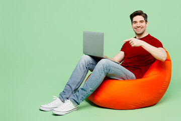 Full body smiling happy young IT man he wearing red t-shirt casual clothes sit in bag chair hold use work point on laptop pc computer isolated on plain pastel light green background studio portrait.