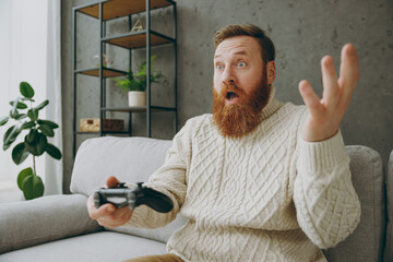 Young confused man in casual clothes hold in hand play pc game with joystick console sit on grey sofa couch stay home hotel flat rest relax spend free spare time in living room indoor Lounge concept © ViDi Studio