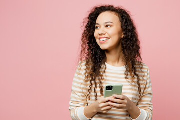 Young smiling happy woman of African American ethnicity she wears light casual clothes hold in hand use mobile cell phone look aside on area isolated on plain pastel pink background studio portrait.