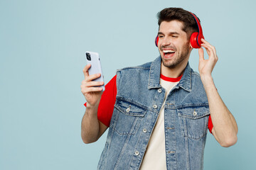 Young cheerful man wear denim vest red t-shirt casual clothes headphones listen music use hold...