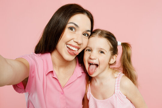 Close up woman wear casual clothes with child kid girl 6-7 years old. Mother daughter do selfie shot pov on mobile cell phone show tongue isolated on plain pink background. Family parent day concept.