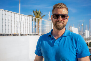 Portrait of handsome smiling stylish hipster model.Modern man dressed in blue shirt. Fashion male posing near skyscraper on the street background in sunglasses. Outdoors at sunset