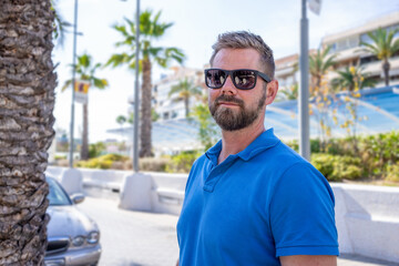 Fototapeta premium Portrait of handsome smiling stylish hipster model.Modern man dressed in blue shirt. Fashion male posing near skyscraper on the street background in sunglasses. Outdoors at sunset