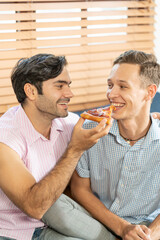 Cheerful gay couple eating pizza. Gay couple sitting on sofa at home,  eating one slice of pizza. Homosexual relationship concept.