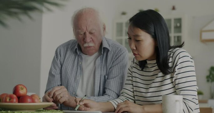 Caring nurse helping old man with paperwork and using smartphone in kitchen at home. Caregiving service and senior people concept.