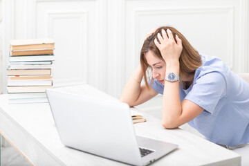 Tired and bored female student reading book and preparing for exams at university. Stack of books and laptop on the desk. Online education and learning from home concept