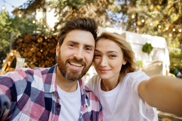 Fototapeta premium A lovely couple are sitting near the travel trailer, taking a selfie, laughing having fun, enjoying their vacation. Portrait of in love enjoying a sunny day outside the city, camera view.