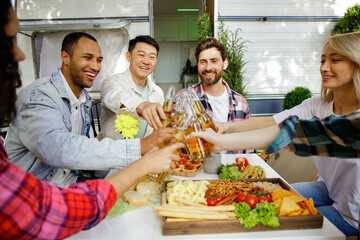 A cheerful multinational company is having fun on a picnic. Multisex friends sitting at the table outdoors celebrating eating and drinking drinks.