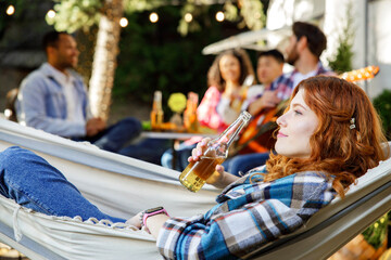 A redhaired girl lies in a hammock in nature. A young woman is relaxing near a tourist trailer in the company of friends, having fun.