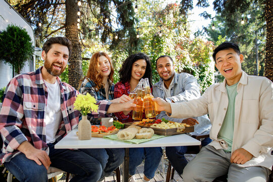A cheerful multinational company is having fun on a picnic. Multisex friends sitting at the table outdoors celebrating eating and drinking drinks. Look at the camera.