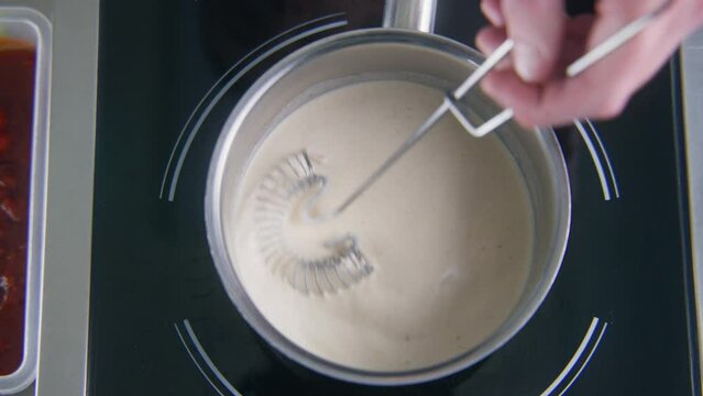 Close up shot of chef cook mixing cream sauce by spiral whisk in stewpan on electric stove. Cook prepares food for guests. Process of making delicious dishes. Work on restaurant kitchen. Slow motion.