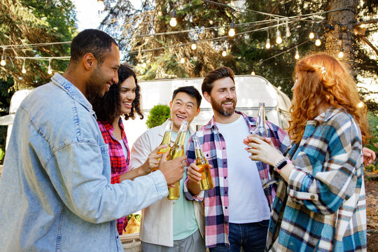 A cheerful multinational company is having fun on a picnic. Multisex friends sitting at the table outdoors celebrating eating and drinking drinks.