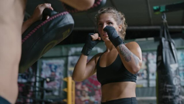Female muay thai fighter training with coach in gym wearing black boxing bandages and athletic clothing. Caucasian woman kickboxer working out punches and kicks with partner