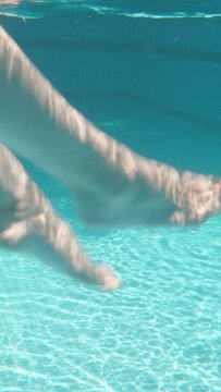 Blue swimming pool, underwater shooting, sunny day. young woman swinging her legs underwater Vertical video.