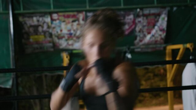 Woman fighter working on her punches in gym ring looking at camera, posters in background. Portrait of female boxer in black sportswear and boxing bandages on wrists and fists training alone