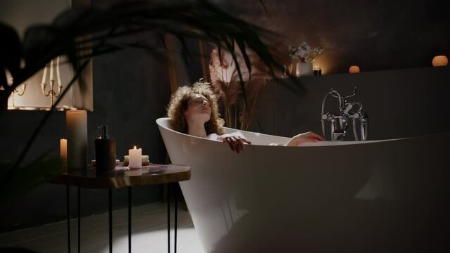 Curly woman enjoying a warm foam bath after a hard day at work. A girl relaxes in a bath and enjoys the smell of aromatic candles placed around.