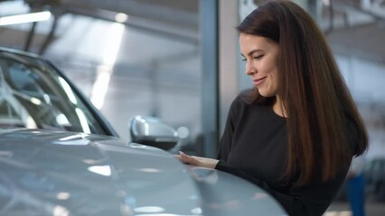 Portrait of young beautiful Caucasian woman touching car hood smiling choosing vehicle in dealership. Confident slim charming buyer making decision purchasing new automobile indoors