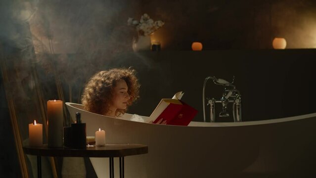 A curly-haired woman lies in a bath and leafs through a book with a red cover. A young woman takes a bath before going to bed and reads her favorite book.